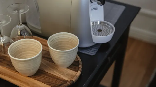 Två keramikkoppar står på en träbricka bredvid en espressomaskin, placerad på ett mörkt bord med två upp-och-nedvända vinglas i närheten.