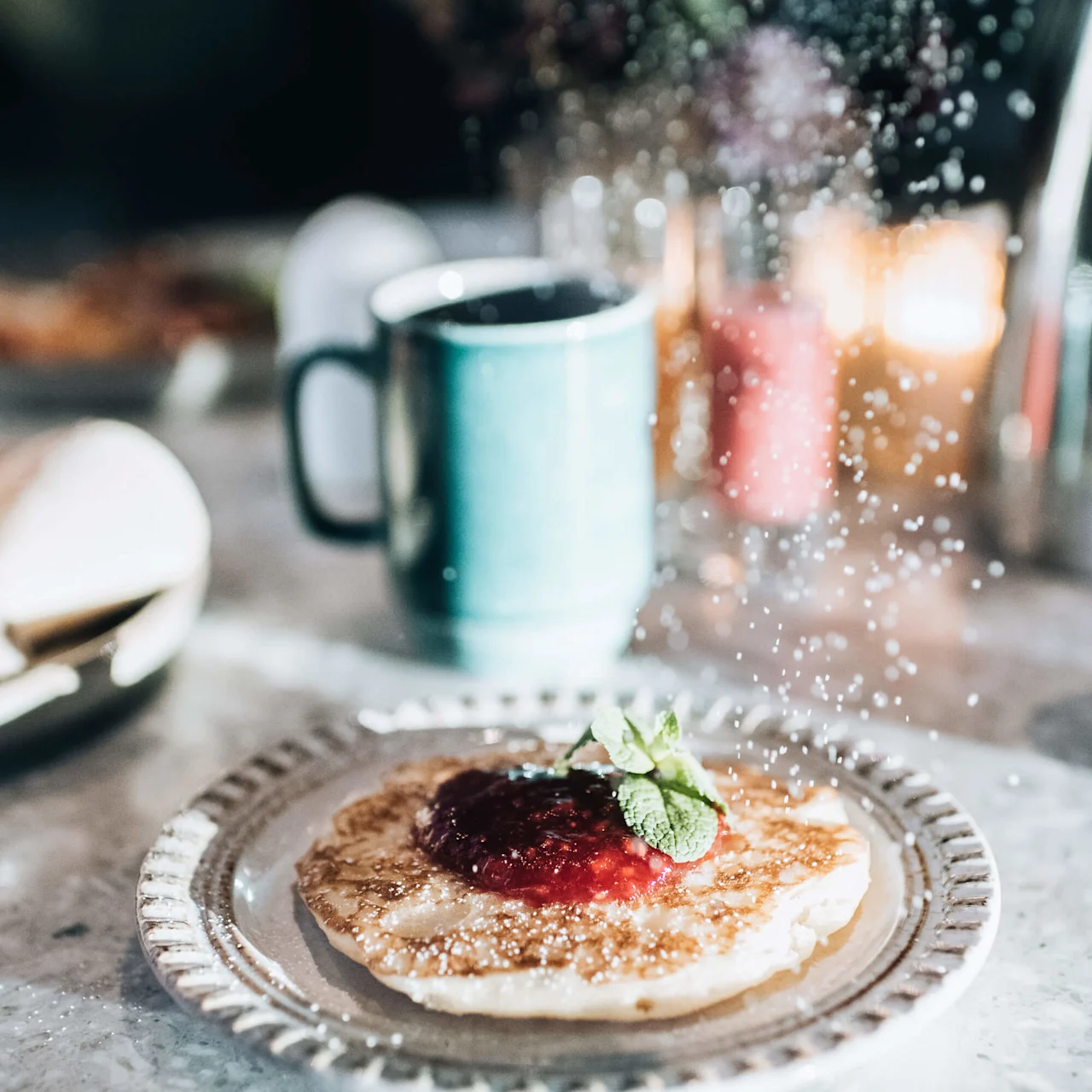 En pannkaka på en tallrik beströs med florsocker, toppad med jordgubbssylt och mynta, på ett bord med en mugg i bakgrunden.