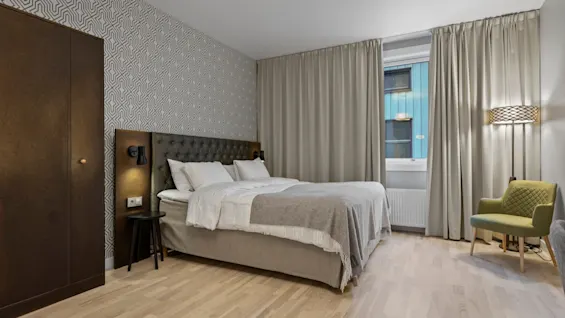 Ett inbjudande hotellrum med en stor, bekväm säng, ljust trägolv och en stilfull fåtölj bredvid ett fönster med gardiner.