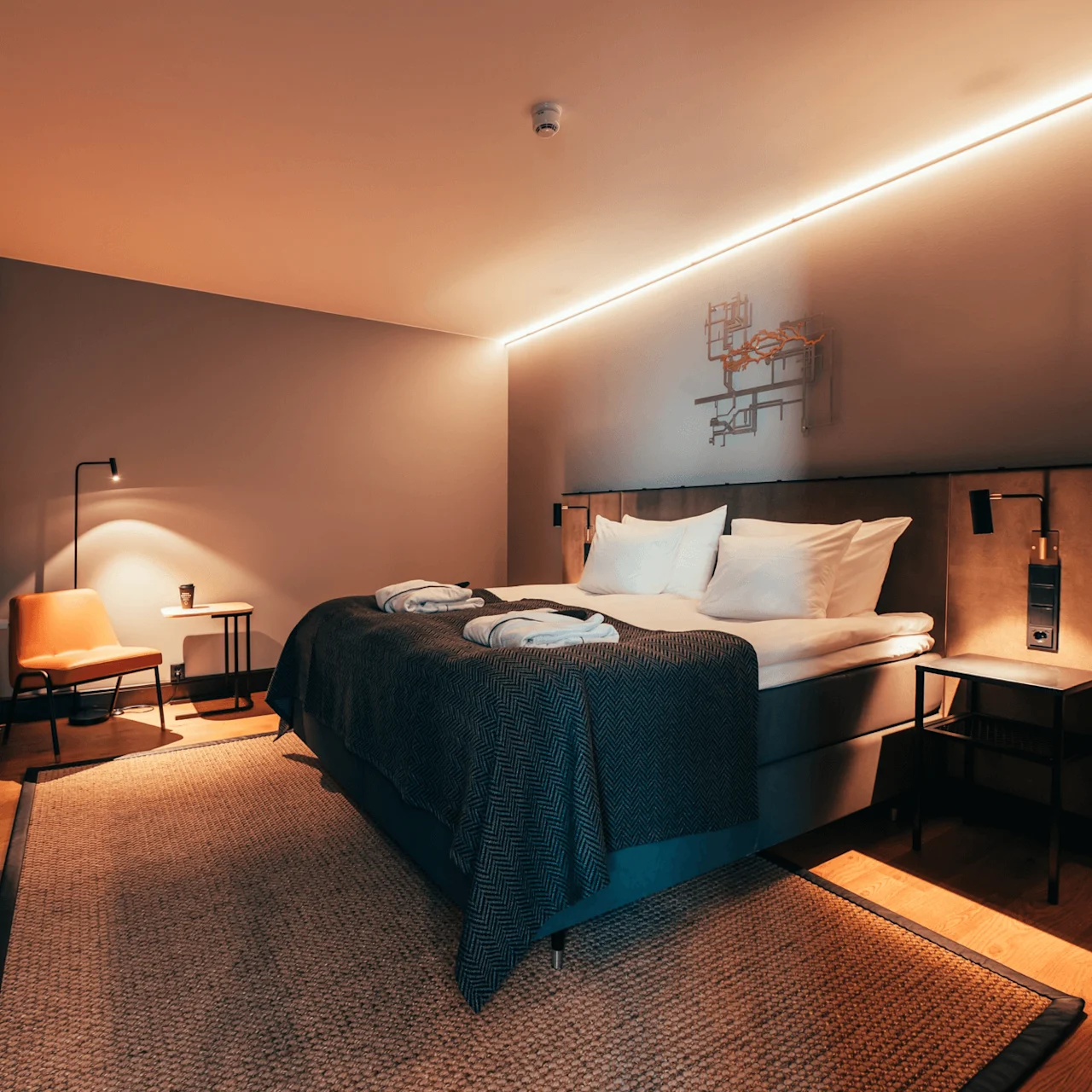 En pent oppredd seng med mørke tepper står i et moderne hotellrom. Myk belysning fremhever en lenestol, sidebord og abstrakt veggkunst. Et teksturert teppe dekker tregulvet.