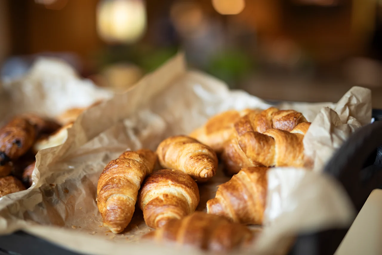 Nystekte croissanter hviler på krøllet bakepapir, plassert i en kurv i en koselig kaféinnstilling. Varm belysning fremhever de gyldenbrune fargetonene til bakverkene. Ingen tekst er synlig.
