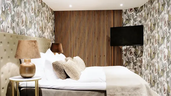 A bed with white pillows and textured cushions stands against a tropical-patterned wallpaper, flanked by two brown lamps on tables in a cozy, warmly lit room.
