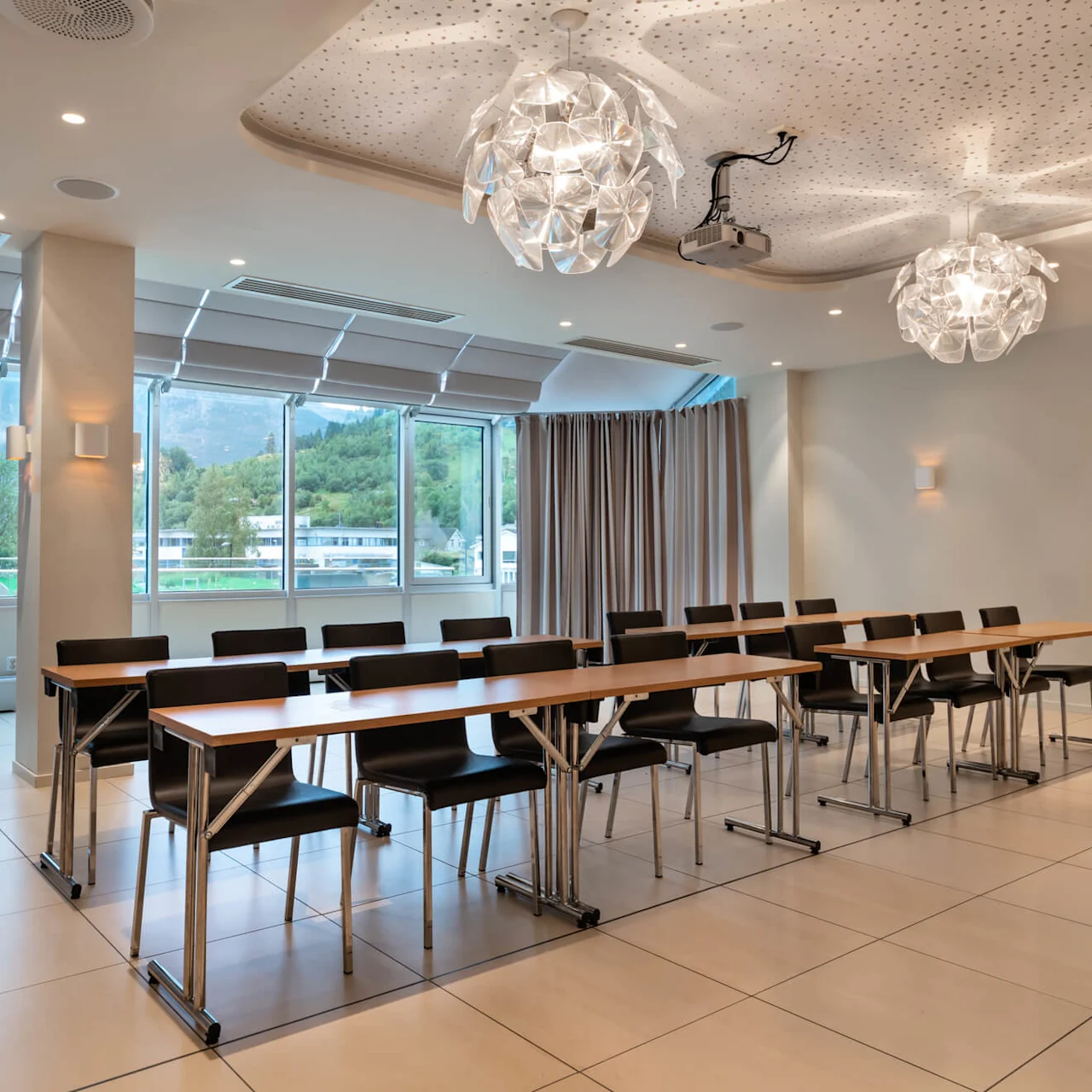 Rader med svarta stolar och träbord är arrangerade i ett modernt konferensrum med stora fönster och eleganta taklampor, med utsikt över ett grönt, trädbevuxet landskap.