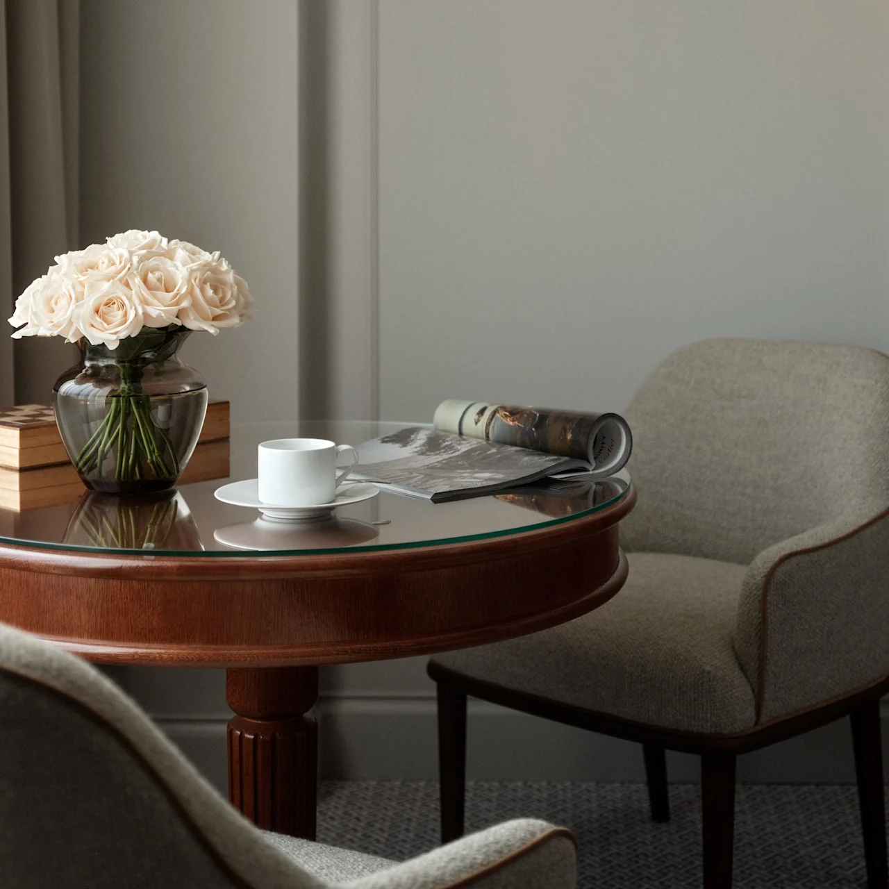 A round wooden table holds a vase of white roses, an open magazine, and a coffee cup, surrounded by two upholstered chairs in an elegantly decorated room.