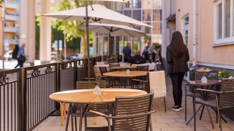 An outdoor cafe seating area with empty tables and sun umbrellas on a sunny day, people in the background.