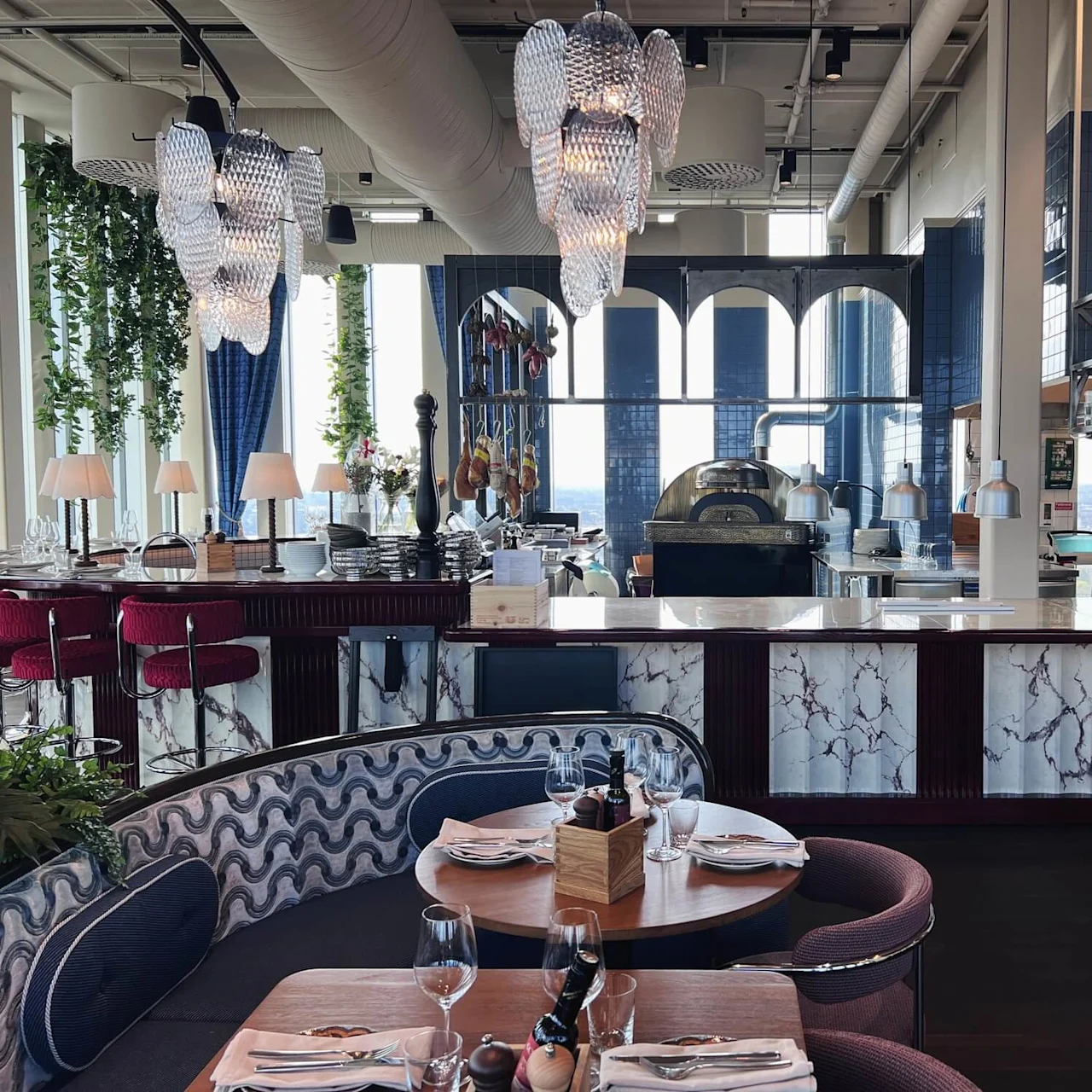 En stilfuld restaurantindretning har borde med borddækning. Lysekroner hænger ovenover, og marmorborde omkranser det åbne køkken, omgivet af røde barstole og dekorative planter.