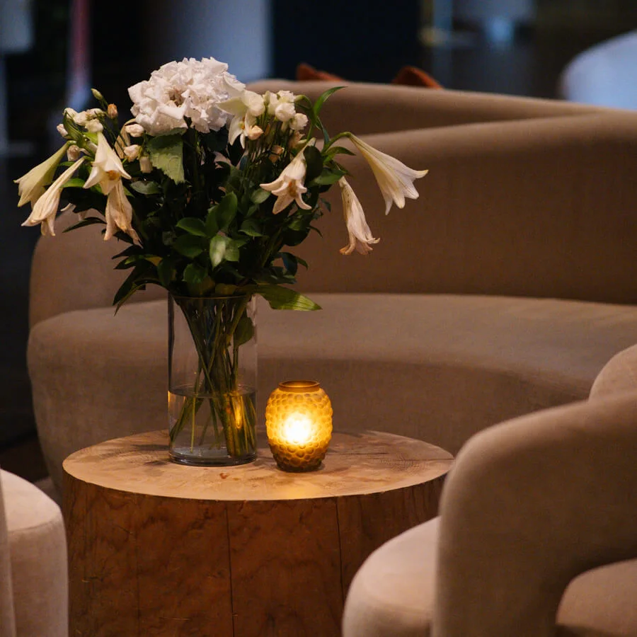 Vase with white flowers sits on a wooden table, accompanied by a lit candle, surrounded by beige sofas in a dimly lit room.