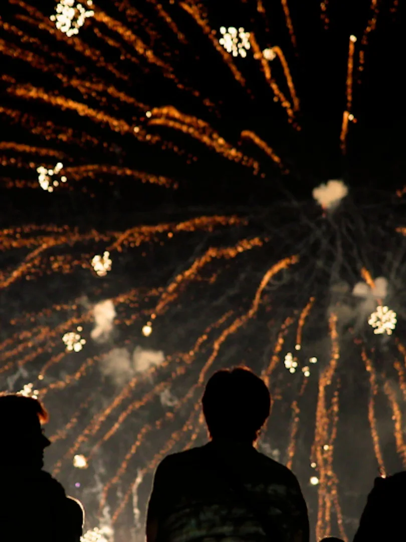 Silhouettes of people watching a vibrant display of orange and gold fireworks exploding in the night sky.
