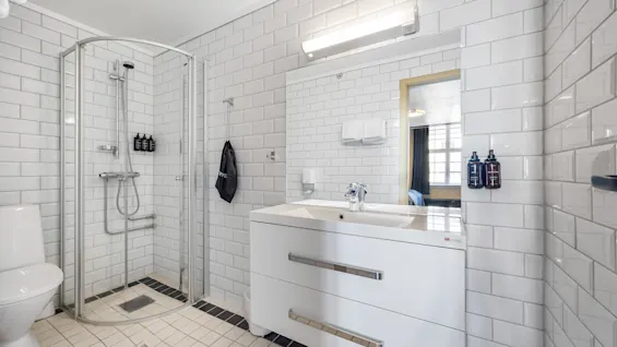 Lyst og moderne hotellbaderom med hvite fliser, dusjkabinett og en stor servantskap med speil.