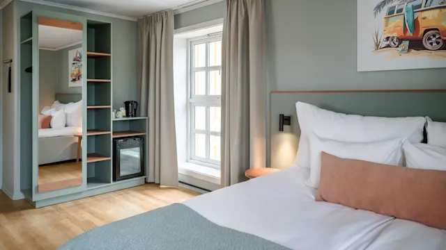 En pent oppredd seng med hvite puter og en rosa pute står i et mykt opplyst rom. Ved siden av står en trehylle med et minikjøleskap og vannkoker ved et vindu.