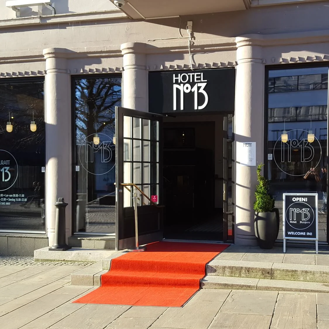 En bygning med søyler har skiltet "HOTEL N°13" over en dør, flankert av vinduer med "RESTAURANT N°13." En rød løper leder til inngangen, med et skilt som sier "OPEN! N°13 Hotel. Burgers. WELCOME IN!"