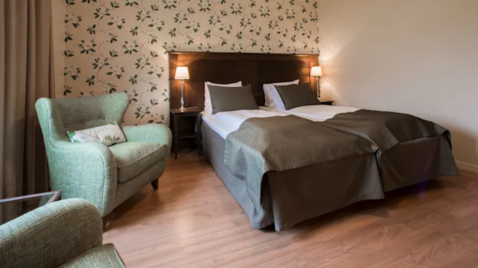 A neatly made bed with grey linens in a room with floral wallpaper, wooden floor, and armchairs.