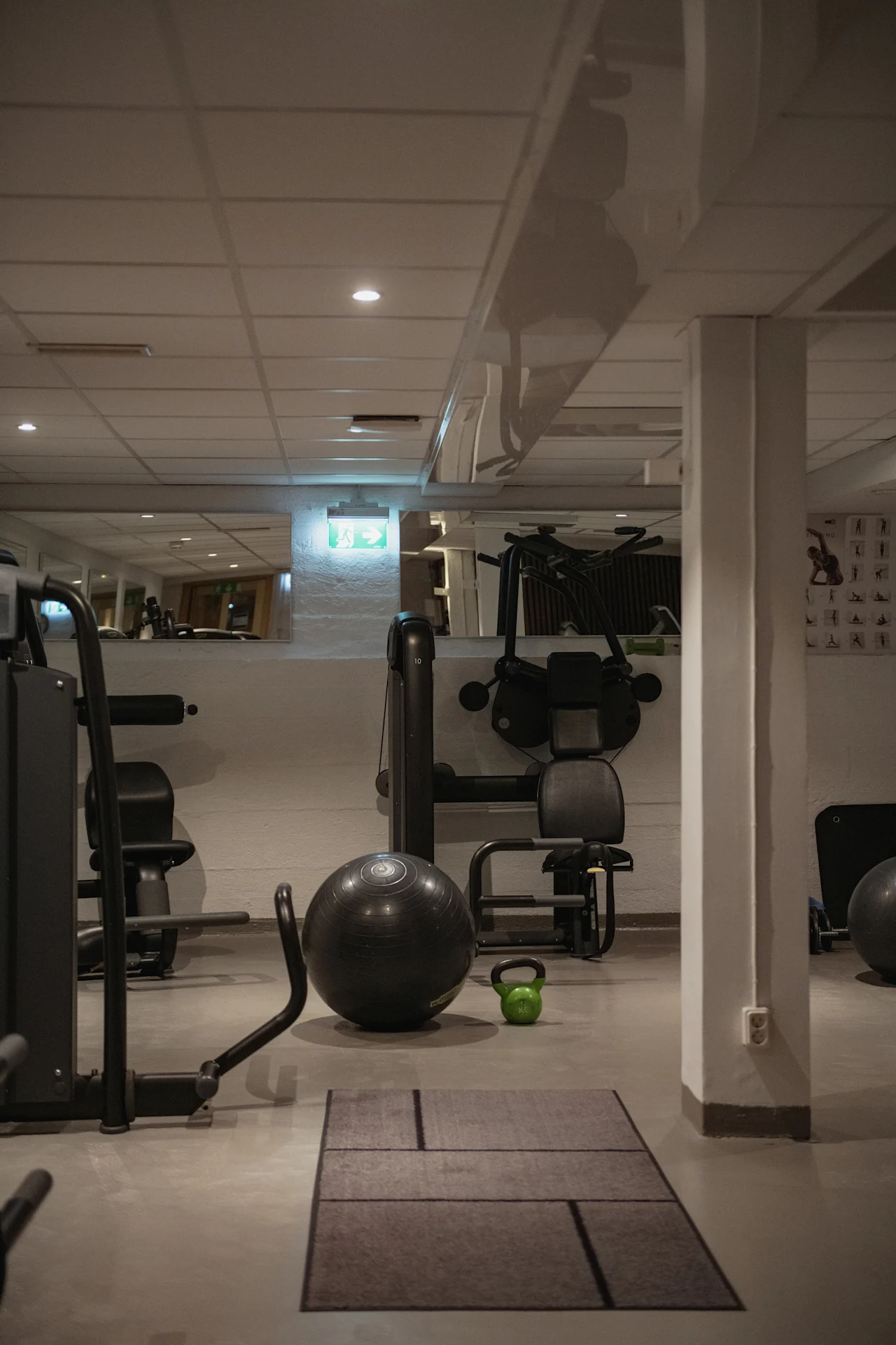 Et rolig treningsstudio med diverse treningsutstyr som en stor svart stabilitetsball, en grønn kettlebell og treningsmaskiner. Et EXIT-skilt viser veien.