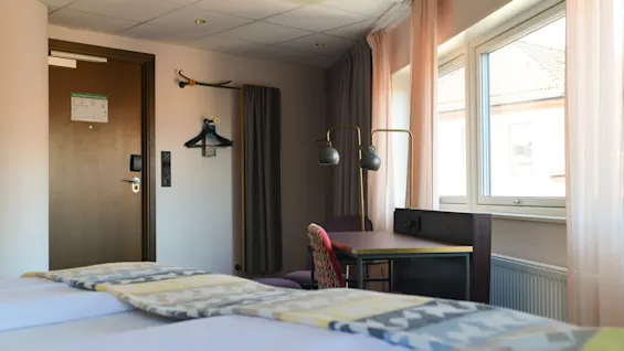 En seng med mønstrede tæpper befinder sig i et hotelværelse. Ved siden af et skrivebord med to lamper ved et vindue. En dør med kroge og bøjler ses.