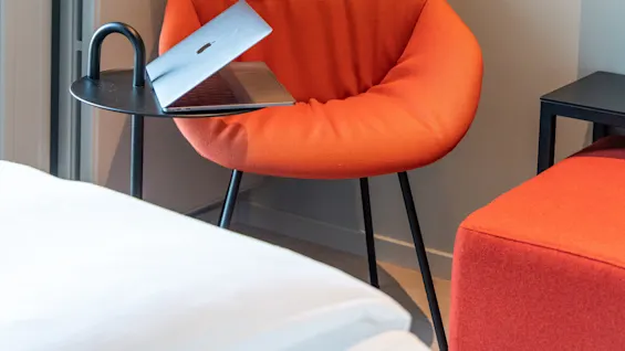 An orange chair holds a partially closed laptop, beside a small black side table. A matching wall lamp hangs above, in a minimalist room.