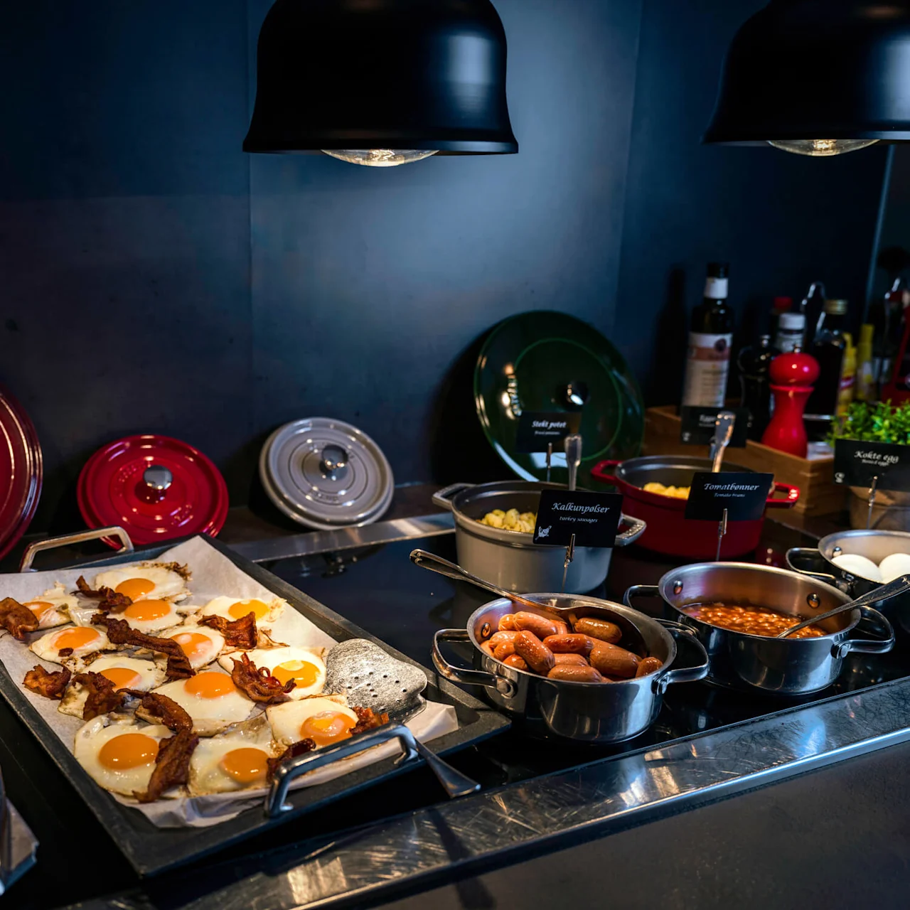 Metal serving trays display fried eggs, bacon, sausages, and beans, under warming lights. Pancakes and cookware with labels "Söt potet," "Kalkunspiker," "Tomatbönner," "Kokte egg" complete the breakfast buffet setup. 