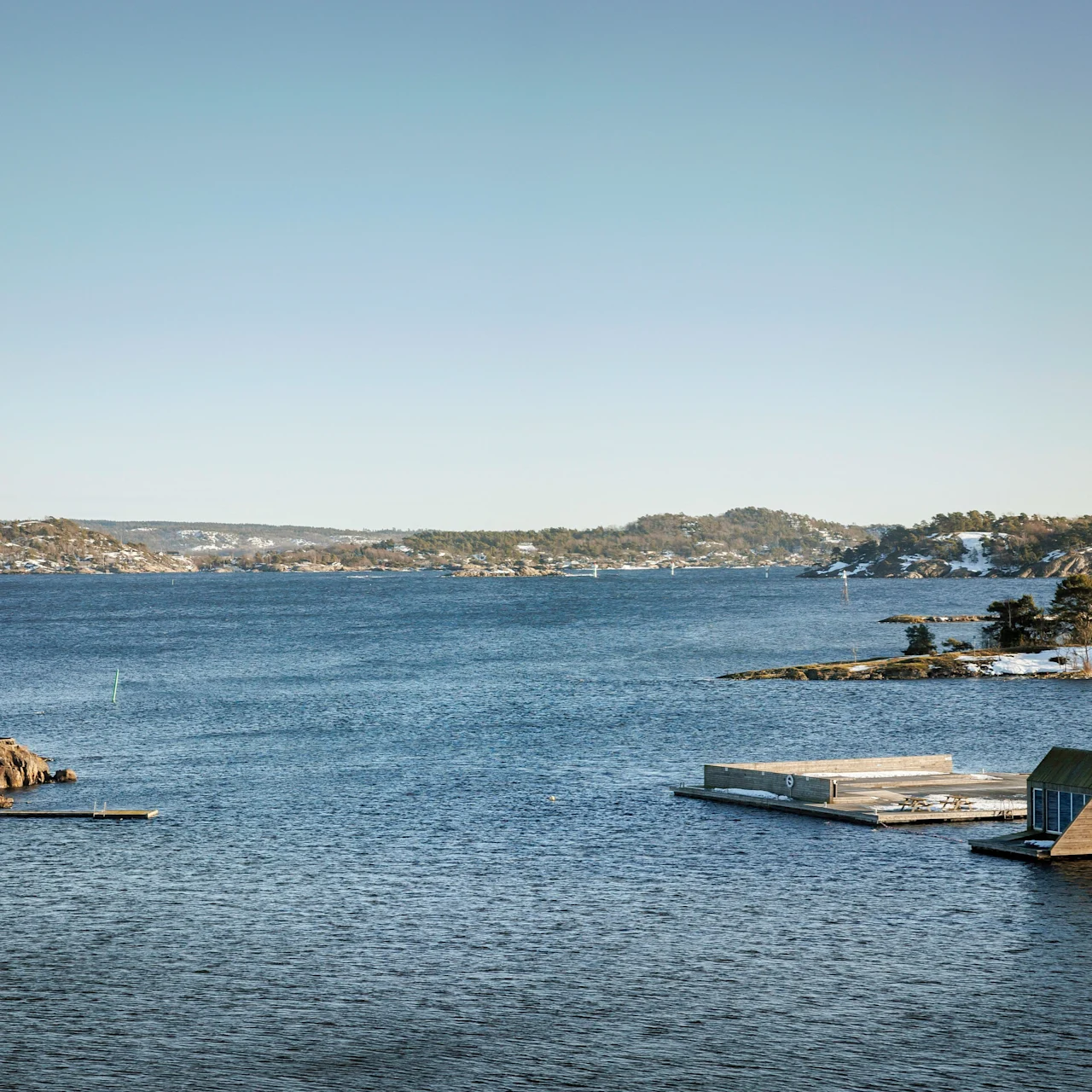 Lighthouse standing on rocky shore, overlooking calm blue water. Wooden docks float nearby, surrounded by an island landscape under a clear sky.