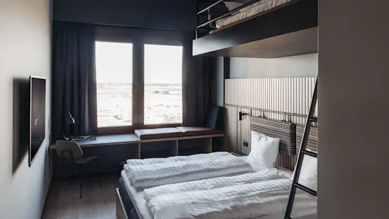 A modern hotel room features a double bed and loft, with a ladder leading to the upper level. A large window overlooks a construction site. A desk and chair sit nearby.