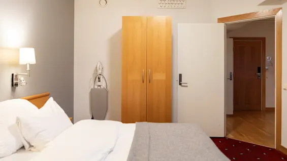 Et koselig hotellrom med en pent oppredd seng, en tregarderobe og et strykebrett, med døren litt på gløtt.