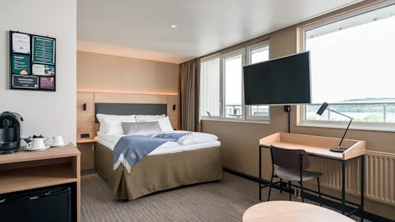 A neatly made bed with white linens and a blue throw sits in a modern hotel room, beside a windowsill desk and wall-mounted TV. A coffee machine and mini fridge are in the foreground.