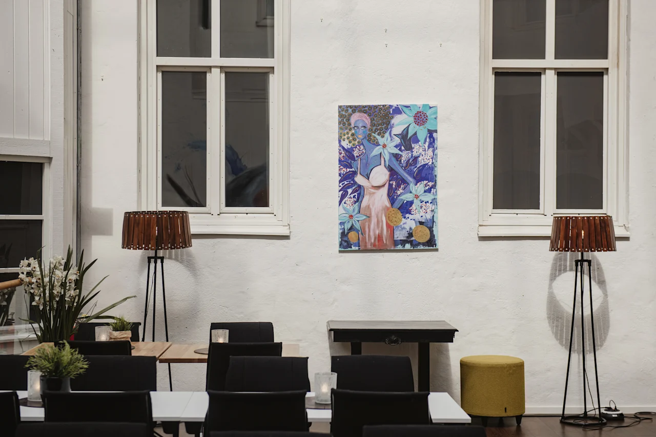 Två höga lampor ramar in en färgglad målning på en vit vägg mellan två fönster. Nedanför står bord och stolar i en stilren, modern inomhusmiljö.