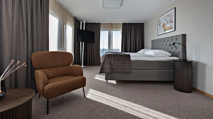 En brun lenestol står på et teppebelagt gulv ved siden av en moderne seng, med høye gardiner og mykt lys som fyller rommet.