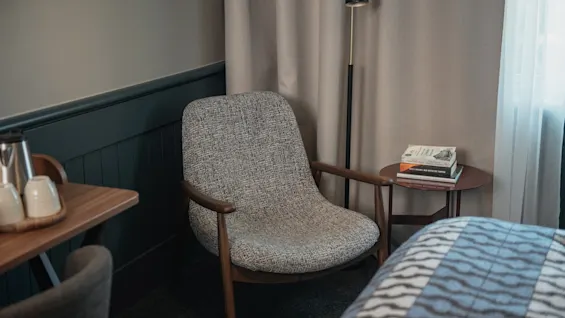 Ett mysigt hotellrumshörn med en bekväm fåtölj, ett sidobord med böcker och en golvlampa, perfekt för avkoppling.