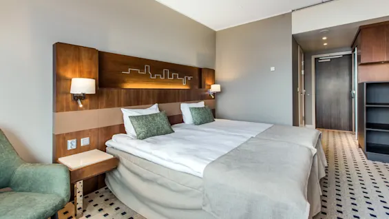 A neatly made hotel bed with green pillows rests against a wooden headboard with skyline design, beside a green armchair and wooden furniture in a modern, softly lit room.