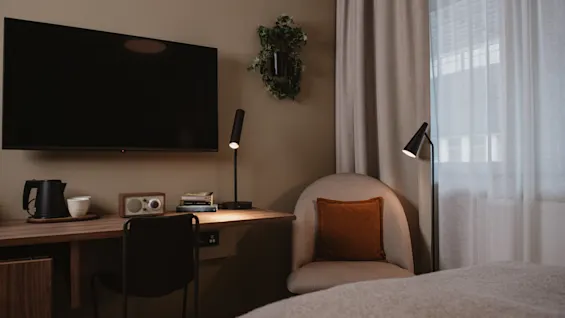 TV mounted on a wall above a desk with a kettle, cup, and radio. A chair sits nearby, with a floor lamp illuminating the area. Curtains cover a window.