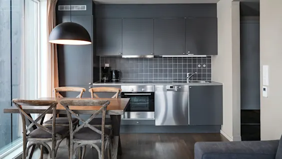 Dark gray modern kitchen with a stainless steel oven and dishwasher, located beneath cabinets and next to a wooden dining table. A large window provides natural light.