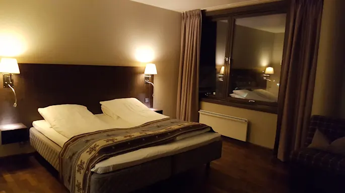 A neatly made bed with white linens is in a dimly lit hotel room, flanked by two wall-mounted lamps. A large window with closed curtains faces the bed.