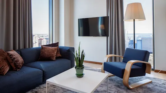 Et moderne opholdsstue har en blå sofa og lænestol placeret omkring et hvidt sofabord med en plante. En fladskærms-TV er monteret på væggen ved siden af store vinduer med udsigt over byen.