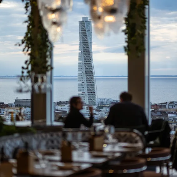 People enjoying a meal or drinks at a restaurant with a panoramic view of a city skyline and a unique twisted skyscraper.