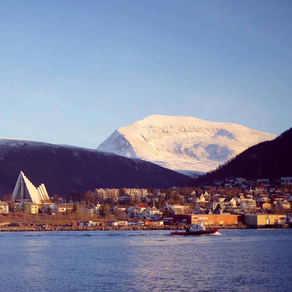 A coastal townscape with a bridge, boats, and a prominent triangular building against snow-covered mountains at sunset.