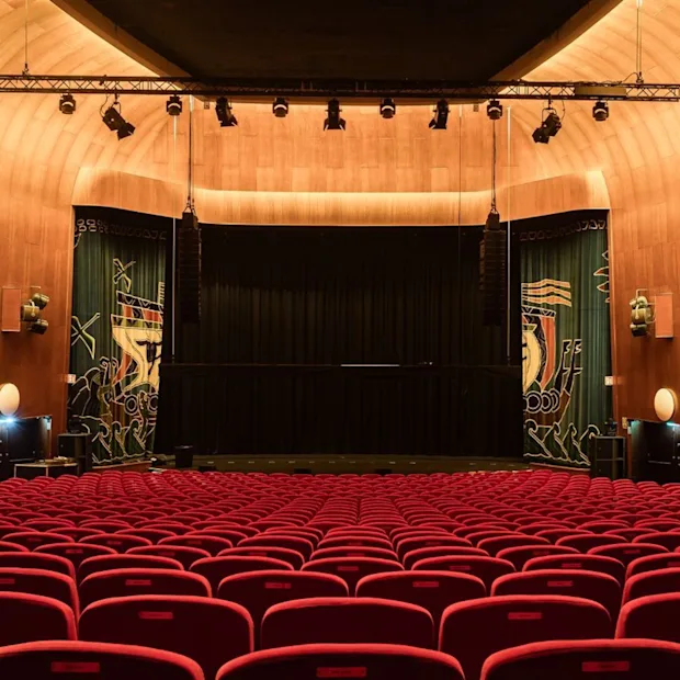 An empty auditorium with rows of red seats facing a stage, ready for an event.