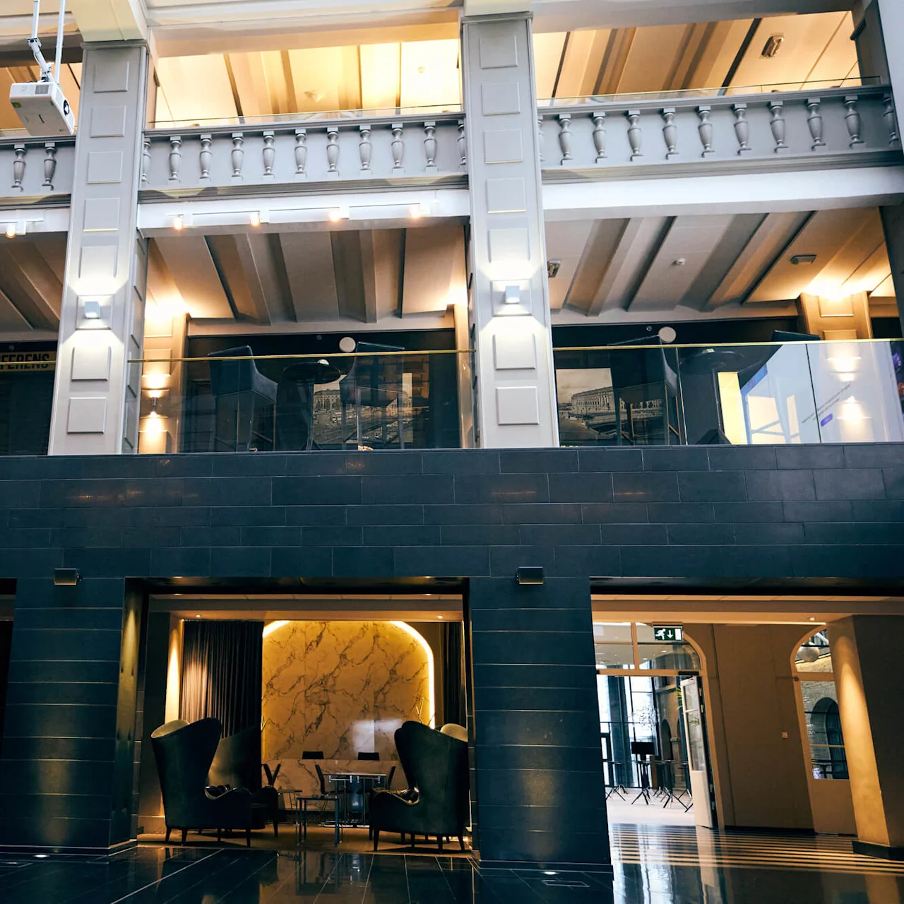 Tilava aula korkeilla katoilla, jossa tyylikkäät tuolit ovat aseteltu pöydän ympärille. Yläpuolella lasiparveke valvoo aluetta. Pehmeä valaistus luo lämpimän ja kutsuvan tunnelman.