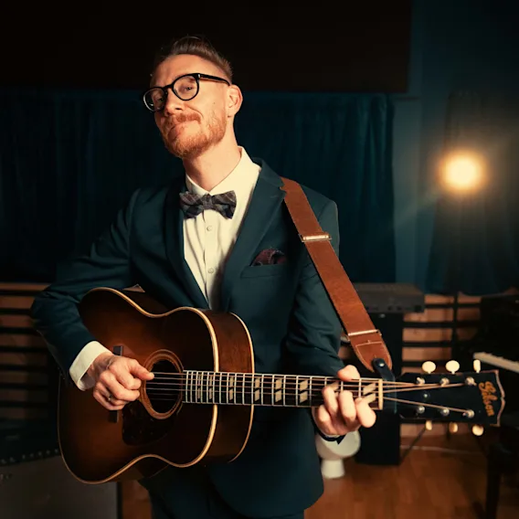 En person i dress og sløyfe spiller akustisk gitar på en svakt opplyst scene med en spotlight i bakgrunnen.