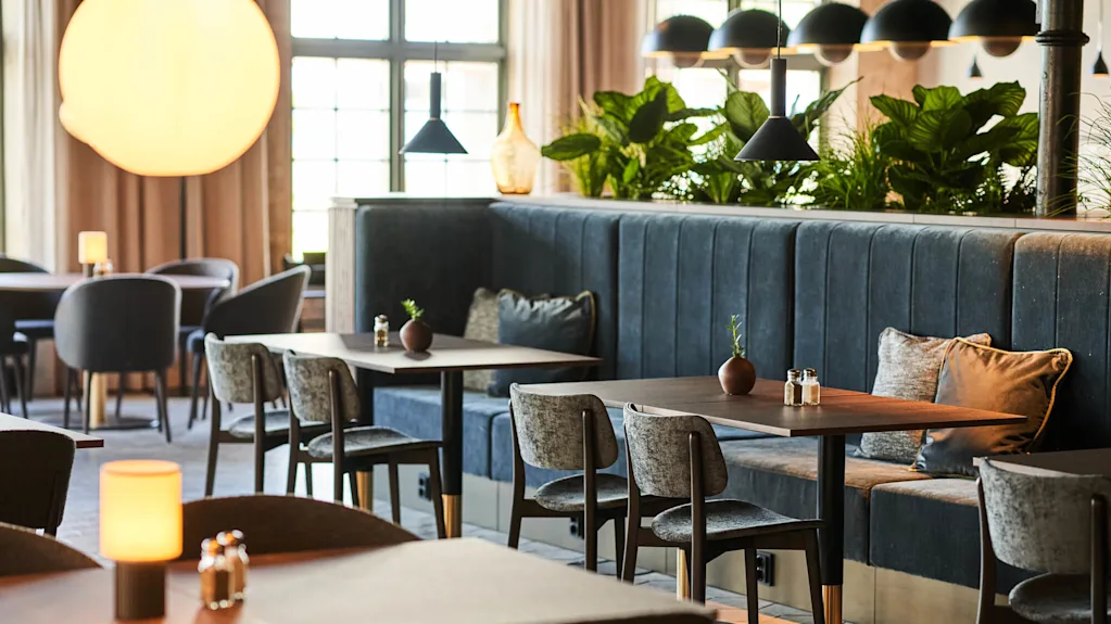 Moderna restaurangbord med grå stolar och mörka bänkar under hängande lampor; gröna växter dekorerar avdelaren, och naturligt ljus strömmar in genom stora fönster.