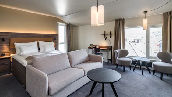 Bed and sofa in a well-lit hotel room; decorated with chairs, tables, and lamps, set against large windows and a desk. Neutral tones create a cozy atmosphere.