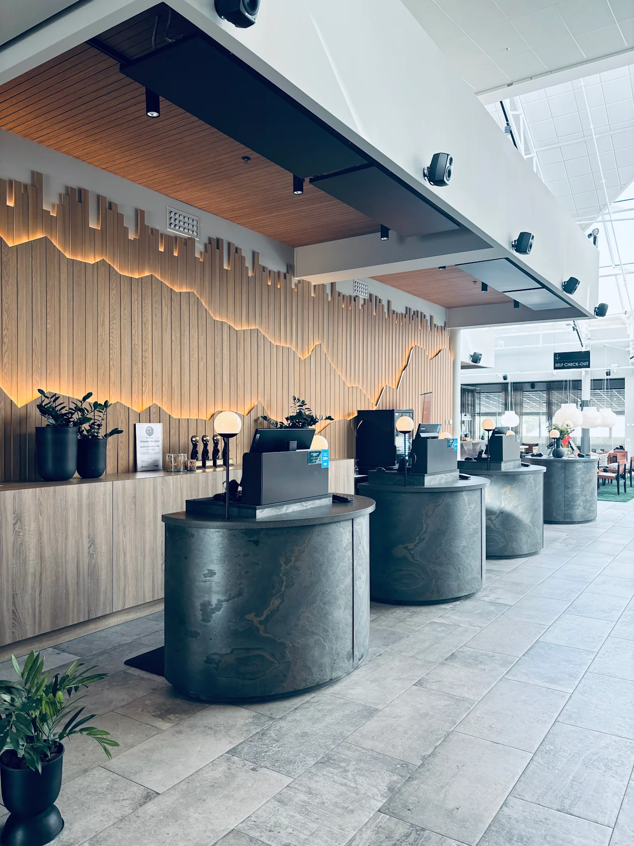 En moderne hotellobby med flere selvbetjente innsjekkingsstasjoner. En dekorativ trevegg med bakgrunnsbelysning gir varme. SELF CHECK-OUT.