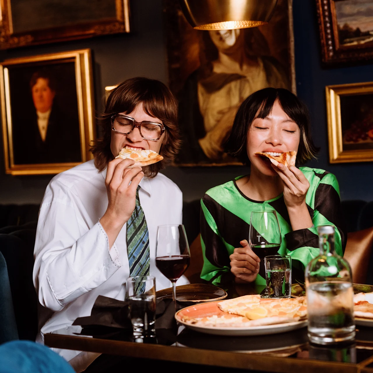Two people are sitting at a table, enjoying slices of pizza. They are in a dimly lit, elegant restaurant with framed paintings on the walls.