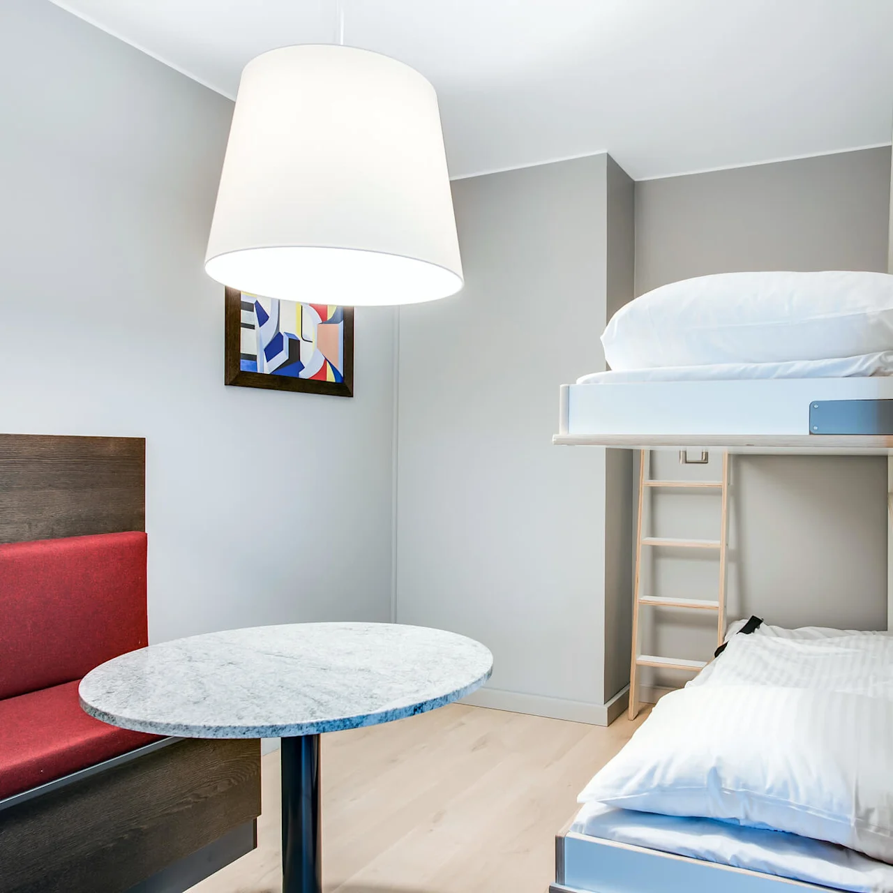 Et moderne rum med køjeseng, rød polstret bænk, marmorbord, abstrakt indrammet maleri og stor loftslampe. Neutrale vægge og trægulv skaber en minimalistisk indretning.