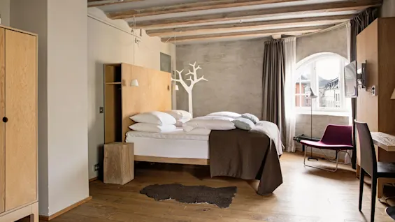 Bed positioned against a concrete wall, white linens, brown blanket; wooden floor. Light streams through an arched window with a view outside. Minimalist furniture includes a chair and desk.