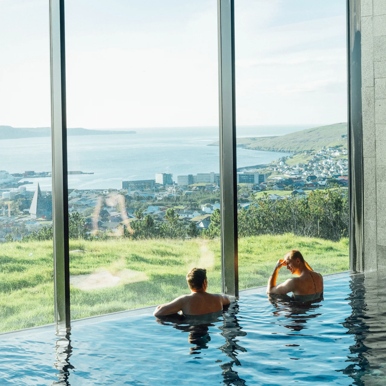 Two people relaxing in an infinity pool with a panoramic view of a coastal town and green landscape.