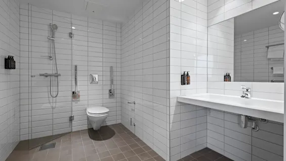 Ett ljust, tillgängligt badrum med vita kaklade väggar, en öppen dusch, en toalett och en rymlig kommod med spegel.