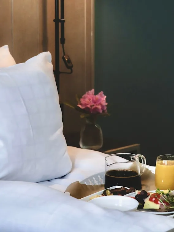 Breakfast tray rests on a bed with white pillows, holding coffee, juice, and a plate of food. A vase with pink flowers sits on a nearby table.