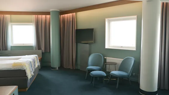 Et soveværelse har en seng og to stole med et lille bord i et hjørne. Vinduer lader naturligt lys ind, mens gardiner og et fjernsyn fuldender det moderne, minimalistiske rum.