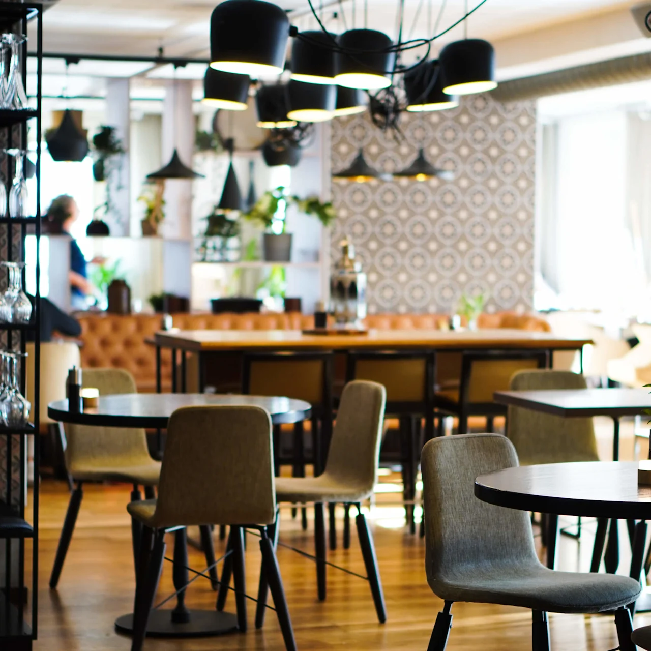 Borde og stole står ubrukte i en moderne kafé, opplyst av pendellamper, med mønstret tapet og tregulv som forsterker den koselige atmosfæren.