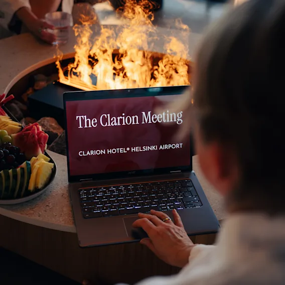 Kylpyläkokous Clarion Hotel Helsinki Airportissa. Henkilö kannettavalla nuotion ääressä hedelmien kera, näyttäen 'The Clarion Meeting'.