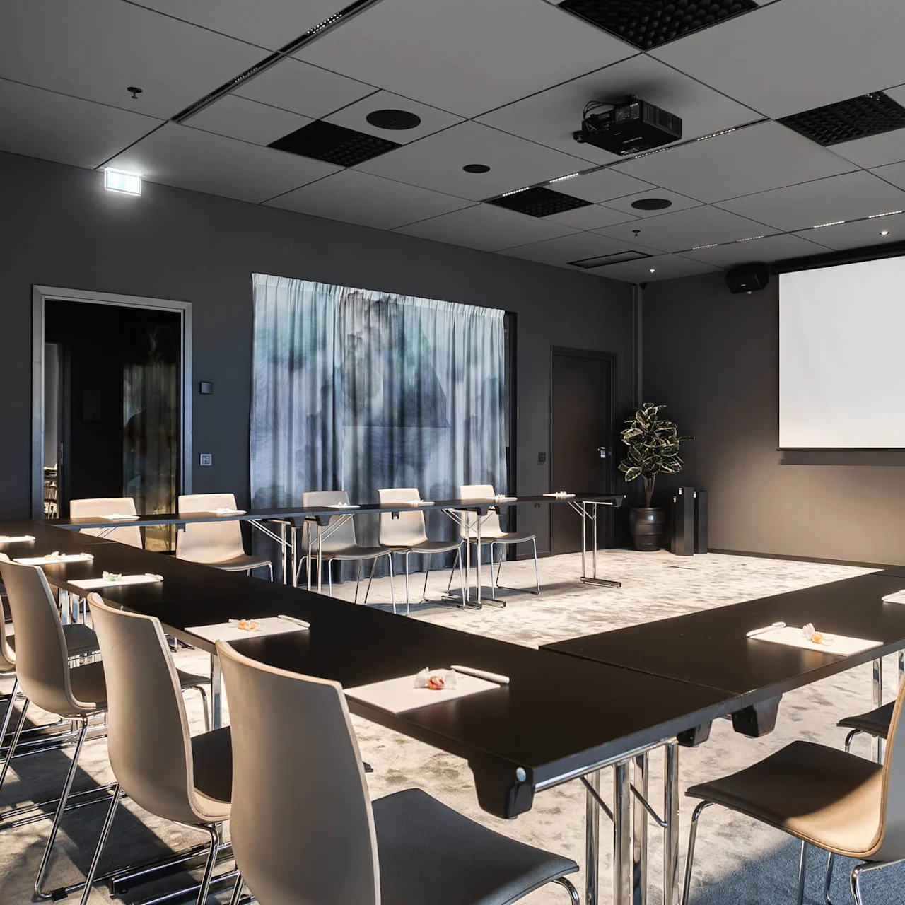 Konferensrum med U-format bordsarrangemang omgivet av beige stolar, vänd mot en stor tom skärm. En krukväxt accentuerar det moderna inredda rummet med mönstrat tak.